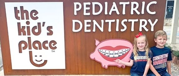 Two smiling kids sitting in front of The Kid’s Place Pediatric Dentistry sign and pig mascot in Boerne, TX