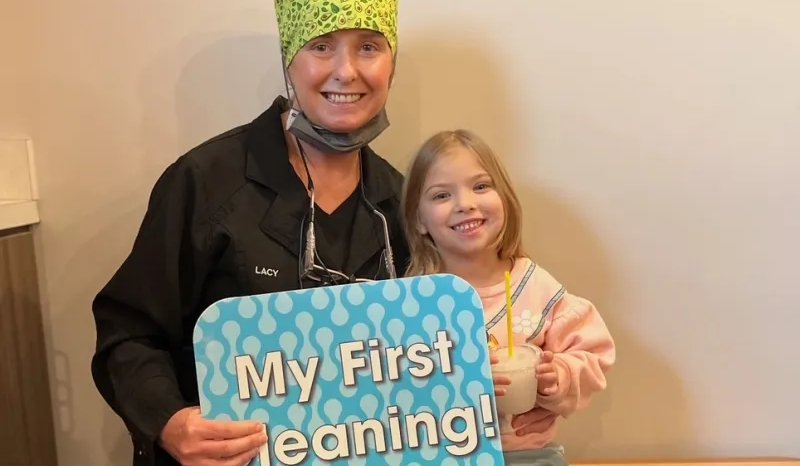 Smiling little girl holding a My First Cleaning sign with a team member at The Kid’s Place in Boerne, TX